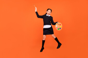 Fototapeta na wymiar Photo of painter cute schoolgirl jump hold palette brush wear uniform isolated orange color background