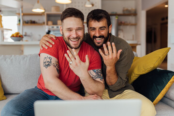 Young happy gay couple in love just got married so they have a video call with their friends and family to spread the joy. The couple showing their wedding rings. - obrazy, fototapety, plakaty