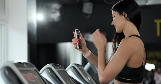A woman takes a selfie on the treadmill in the gym. Attractive girl running on treadmill and taking selfie with smartphone in gym.