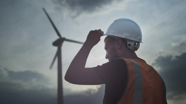A male engineer or repair worker stands next to a wind turbine. Man looks at the propeller and fan blades. White safety construction helmet. Alternative energy sources. Green Fuel Generator