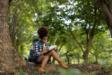 Naklejka na ściany i meble Young women teenager ethnic African American black skin wearing plaid shirt and her backpack sitting at tree base looking down take note in notebook in her hand