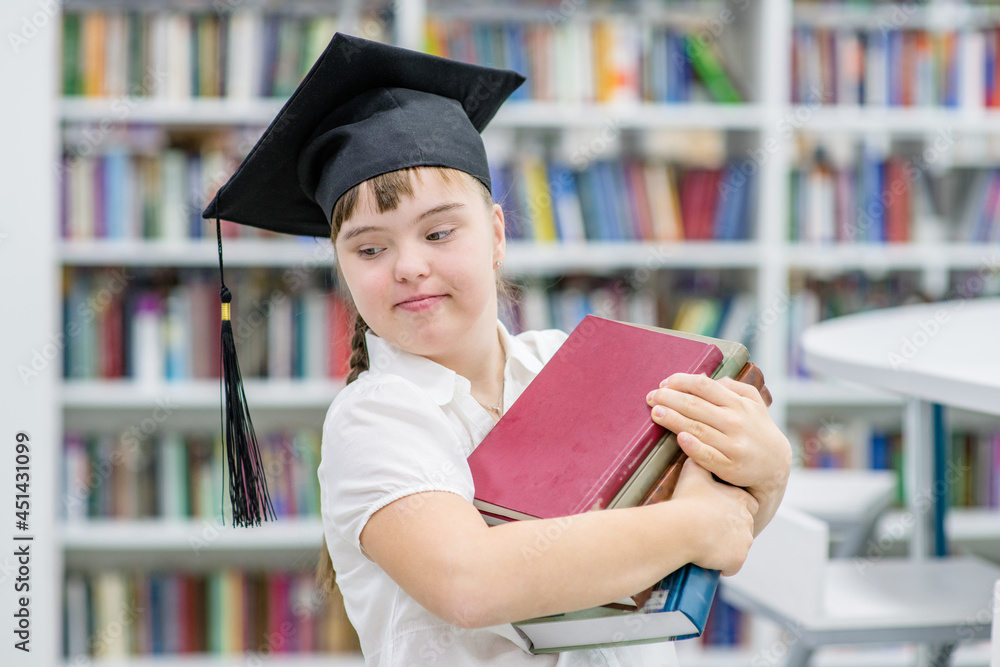 Wall mural happy girl with down syndrome wearing a graduation cap holds books in her hands at a library. empty 