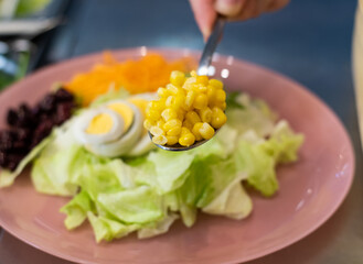 cropped view of woman hand adding corn in pink plate with salad in restaurant. Serving healthy vegetarian salad