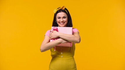 Woman smiling at camera while hugging gift box isolated on yellow.