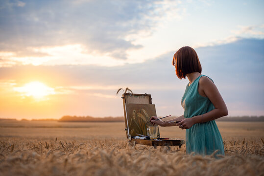 Girl artist drawing a painting in the field of wheat with a setting sun in the background. Beautiful atmospheric shot of a female painter drawing slill life picture with oil inks on canvas