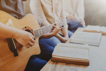 A young man is playing guitar and sings a song from a Christian hymn book with his friends at home,...