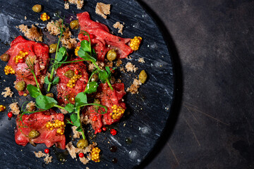 fresh beef carpaccio with capers, mustard and arugula on black board