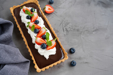 Chocolate cream tart with whipped cream, fresh blueberries and strawberries on top on a dark background. Summer dessert.