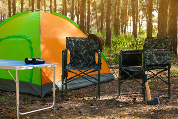 Camouflage chairs with hat, axe and table near camping tent in forest on sunny day