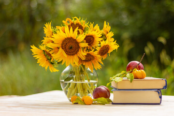 a stack of books and plums next to a glass vase with yellow sunflowers on an old wooden table in...