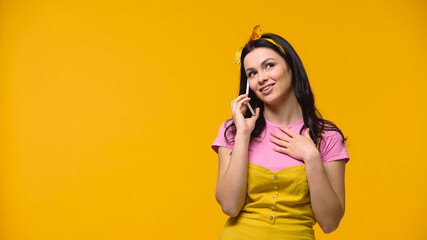 Dreamy woman in headband talking on smartphone isolated on yellow.