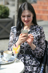 Charming young woman using her smartphone while having cup of coffee. Breakfast on a summer day on terrace in restaurant.