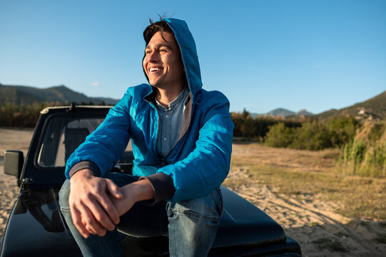 Young smiling man sitting on off-road vehicle with barefoot looking at horizon during sunset. Road trip concept.