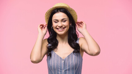 Cheerful woman with closed eyes adjusting sun hat isolated on pink.