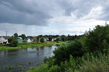 Fototapeta na wymiar Panoramic view of the Tvertsa river. View of the river and wooden houses on the bank. Before the rain, clouds hang over the river.