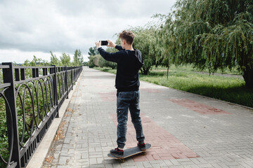 A curly-haired teenager of European appearance in a black hoodie standing on a skateboard in an alley takes a selfie