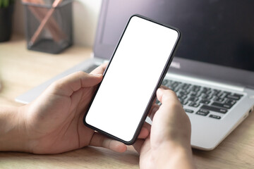 Man's hands holding or using smartphone with two hands in front of working space desk, laptop. Smartphone with white blank on screen, Cellphone mockup.