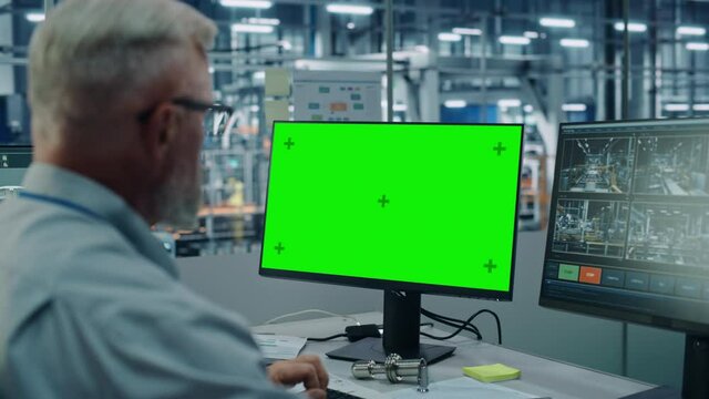 Car Factory Monitoring Office: Male Chief Automotive Engineer Sitting at the Desk Working on Green Screen Chroma Key Computer. Automated Robot Arm Assembly Line Manufacturing Vehicles