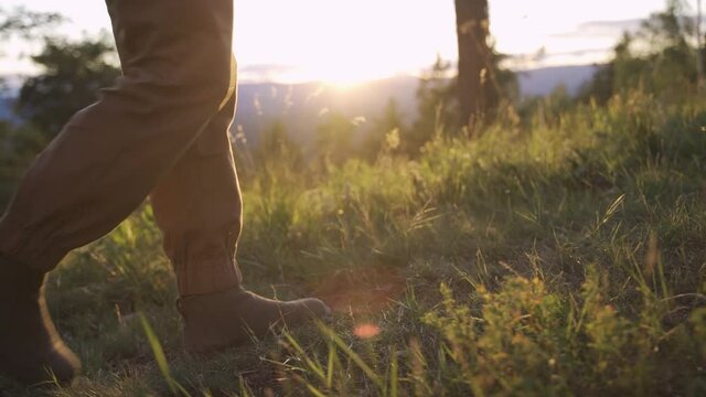 A man in rough boots and hiking pants walks on the grass. It is illuminated by the late sun. Far from home and the big city. Close-up, slow motion, HD.