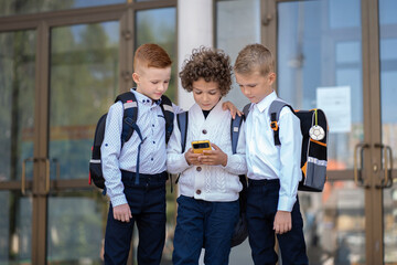 Three little schoolchildren stand at the school and look at the phone