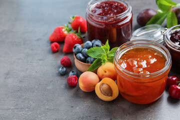 Jars with different jams and fresh fruits on grey table. Space for text