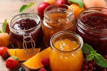 Jars with different jams and fresh fruits on wooden table, closeup