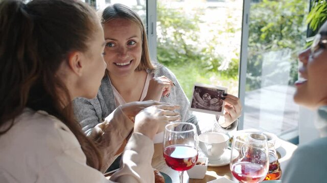Happy future mother is showing sonogram scan image to friends women in cozy summer cafe discussing pregnancy sharing emotions. Friendship and maternity concept.