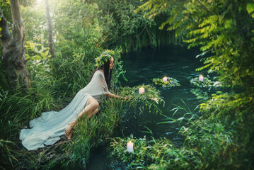 Fabulous mythical natural landscape. Forest fantasy woman sits on shore lake, nymph throws wreath in water. Old Slavic cult ceremony Ivan Kupala. Long white dress. Black hair - obrazy, fototapety, plakaty