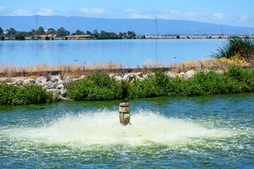 Active working pump aerating and filtering contaminated water in wastewater treatment facility pond to remove pollutants and dissolved gases. Small dam separates oxidation ponds