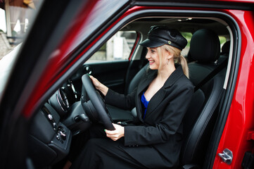 Fototapeta na wymiar Portrait of beautiful blonde sexy fashion woman model in cap and in all black with bright makeup sit and drive red city car.
