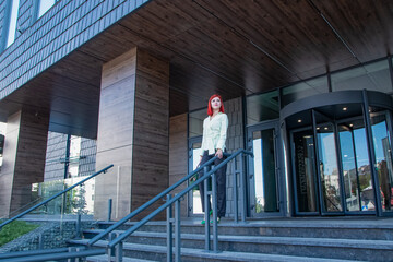 beautiful redhead girl in stylish clothes and a bag near the business building
