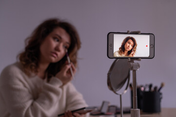 Young caucasian woman is recording an online make-up training video on her mobile. Video blogger girl applies makeup.