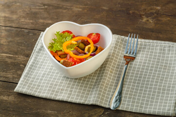 Vegetable salad in a heart shape plate on a wooden table on a napkin.