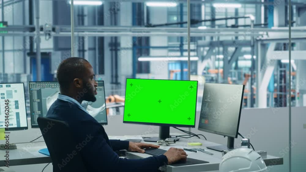 Aufkleber Car Factory Office: Black Engineer Works on a Computer, Two Monitor Screens Show Chroma Key Green Screen and Monitoring of Automated Robot Arm Assembly Line Manufacturing High-Tech Electric Vehicles