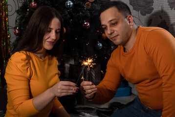 A woman and a man light sparklers and look at them. There is a Christmas tree behind. Selective focus.