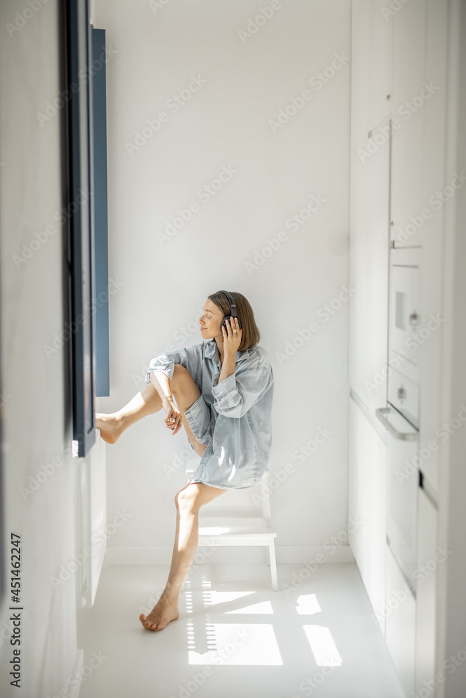 Wall mural Young woman enjoys music in headphones while sitting alone in a bright room at home. Self-isolation and loneliness concept