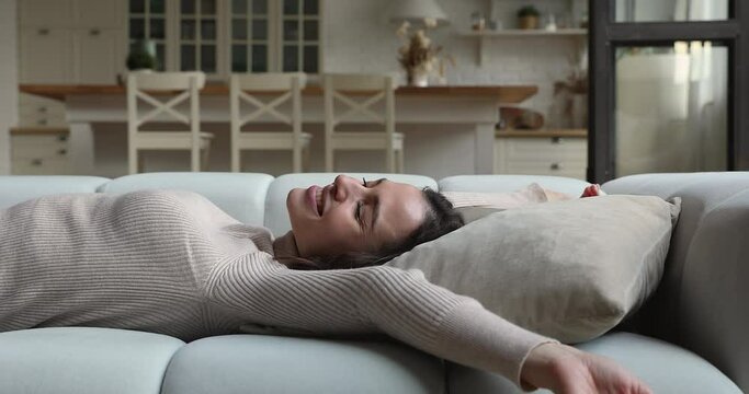 Happy positive young woman fallen on cozy couch, enjoying lazy weekend relaxation time alone at home. Smiling millennial female resting on sofa, raising arms stretching muscles, reposing indoors.