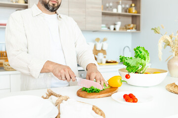 Obraz na płótnie Canvas happy young, handsome, bearded man is standing in the modern kitchen prepares a salad of fresh vegetables, cuts vegetables with a knife on a cutting board, cooking as concept of a man's hobby