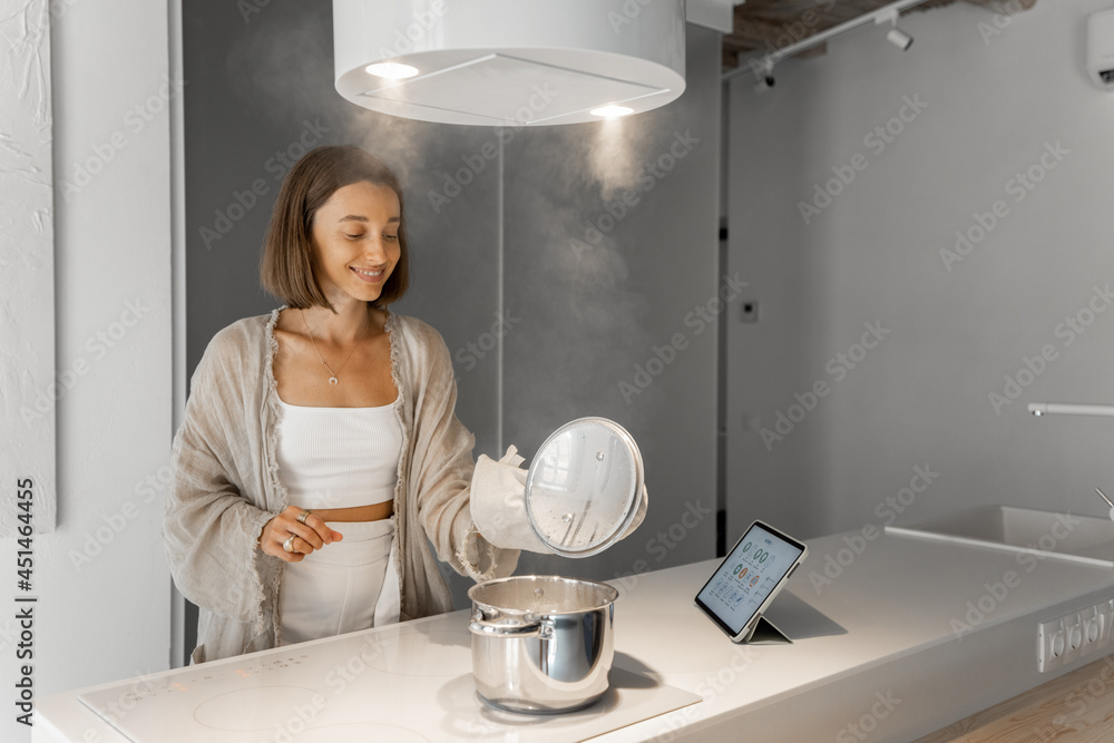 Wall mural Young woman boiling food on electric hob, cooking at modern kitchen at home. Home comfort, lifestyle at home, eating at home