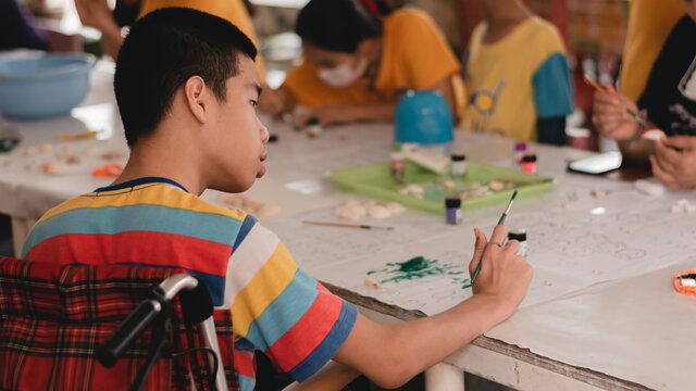 Handicapped Teenager Boy On Wheelchair With Happy Face Doing Art Work With Friends, Lifestyle Of Smart Disabled Kid Learning Activity In Special Children School, Mental Health Classroom Concept.