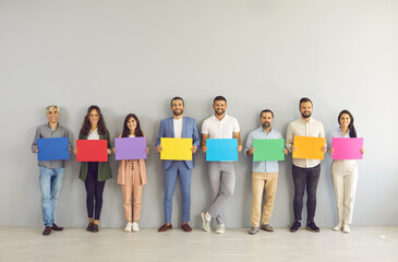 Obraz premium Group portrait of happy smiling young and old business people standing in row by white grey office studio wall holding colorful blue, red, green, yellow, purple, orange, pink mockup paper sign banners
