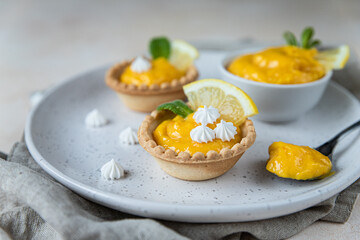 Homemade lemon curd in tartlets with fresh lemon slices, mini meringue and mint on ceramic plate, light background.
