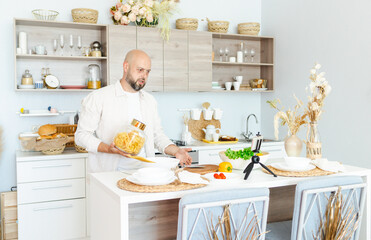 happy young, handsome, bearded man is standing in the modern kitchen with pasta in his hands and vegetables on table, cooking as concept of a man's hobby, records video for food blog