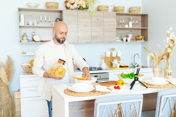 happy young, handsome, bearded man is standing in the modern kitchen with pasta in his hands and vegetables on table, cooking as concept of a man's hobby, records video for food blog