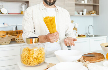happy young, handsome, bearded man is standing in the modern kitchen with pasta in his hands and vegetables on table, cooking as concept of a man's hobby