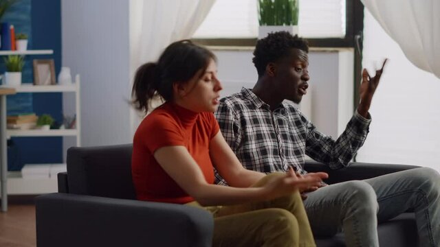 Angry interracial couple fighting on living room sofa. Married mixed race partners having argument about relationship problems and lifestyle. Irritated multi ethnic lovers yelling