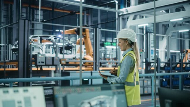Car Factory: Female Automotive Engineer Wearing Hard Hat, Standing, Using Laptop. Monitoring, Control, Equipment Production. Automated Robot Arm Assembly Line Manufacturing Electric Vehicles. Medium