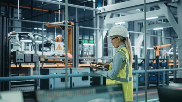 Car Factory: Female Automotive Engineer Wearing Hard Hat, Standing, Using Laptop. Monitoring, Control, Equipment Production. Automated Robot Arm Assembly Line Manufacturing Electric Vehicles. Medium