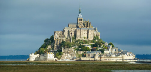 View on french island during low tide illuminated by morning sun with old medieval village and church against misty sky - Mont Saint Michel, France
