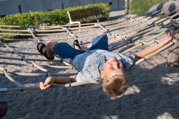 Cheerful blond boy smiling and lying on a rope swing in public park.